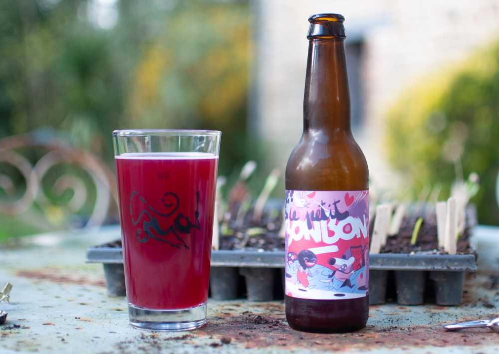 Une photo de bouteille posée sur une table de jardin avec des plants autour, et un verre rempli d'un liquide rougeatre
